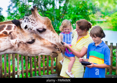 Famille heureuse, jeune mère de deux enfants, rire mignon bébé fille et garçon de l'âge de l'adolescence une girafe d'alimentation lors d'un voyage Banque D'Images