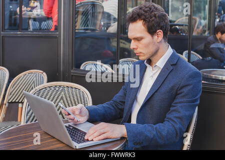 Jeune beau buisnessman en utilisant internet sur téléphone mobile tout en restant assis dans open street cafe Banque D'Images