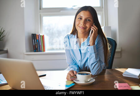 Souriante jeune femme propriétaire d'entreprise avec un tasse de café et téléphone au bureau en bois avec fenêtre derrière son Banque D'Images