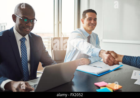 Smiling young Hispanic business man and woman shaking hands à travers une table dans le bureau vu par un smiling African American m Banque D'Images
