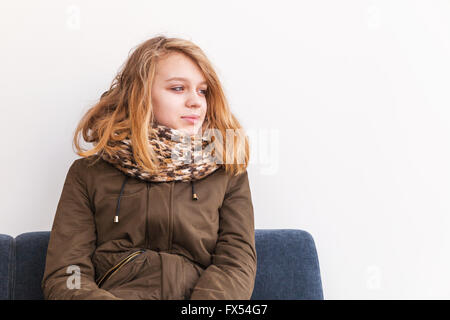 Belle blonde Student graduation dans des vêtements chauds assis sur canapé sur mur blanc Banque D'Images