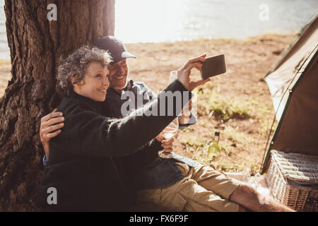 Shot of a senior couple prendre une photo d'eux-mêmes tandis que l'extérieur d'une tente. Mature couple camping près d'un lac en tenant un wit selfies Banque D'Images