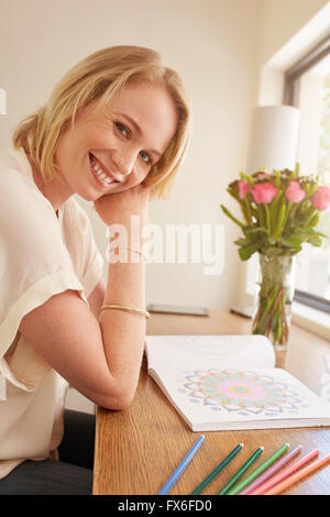 Portrait of happy young woman sitting at a table avec des profils Coloring Book et crayons de couleurs. Young woman relaxing at home Banque D'Images