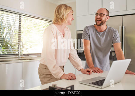 Tourné à l'intérieur d'un couple heureux debout à la cuisine avec un ordinateur portable. L'homme et la femme se regardant et souriant. Banque D'Images