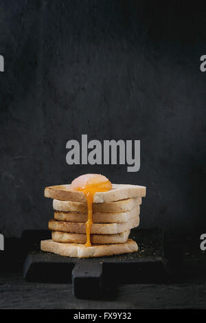 Toasts de pain avec de l'oeuf sucré qui coule sur la planche à découper en bois noir sur fond texturé noir. Banque D'Images