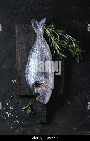 Deux matières prêtes à cuire le poisson dorade au romarin herbes et du sel de mer sur planche à découper en bois sur fond texturé noir. Haut de page vi Banque D'Images