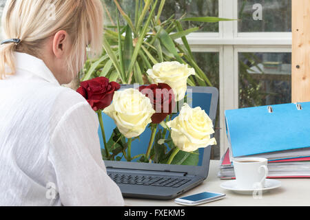 Blonde femme d'âge moyen est assis dans le bureau. À partir de l'ordinateur portable dépasse bouquet de roses rouges et jaunes Banque D'Images