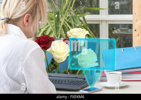 Blonde femme d'âge moyen est assis dans le bureau. À partir de l'ordinateur portable dépasse bouquet de roses rouges et jaunes. Banque D'Images