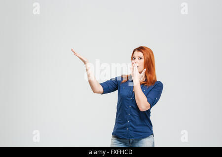 Charmante jeune femme rousse étonnement couvert la bouche avec la main et montrant copyspace over white background Banque D'Images