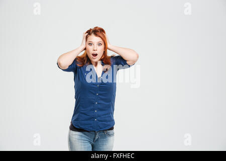 Choqué stupéfié rousse jeune femme avec bouche ouverte debout avec les mains sur la tête sur fond blanc Banque D'Images