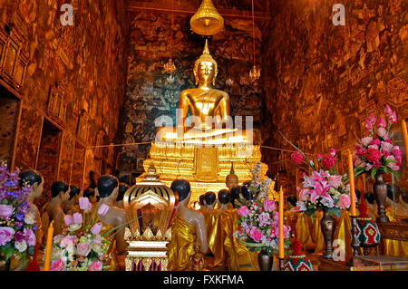 Grande statue de Bouddha en or entourée par des statues de moines en prière le long avec des fleurs et des offrandes à l'intérieur d'un temple bouddhiste Banque D'Images