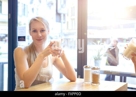 Pretty smiling blonde woman dans un restaurant jouissant d'un latte macchiato avec un ami alors qu'ils s'asseoir et discuter, high key zone de fenêtre Banque D'Images