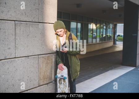 Young male skateboarder urbain leaning against wall lecture textes smartphone Banque D'Images