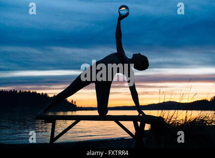 Woman practicing yoga par lac au coucher du soleil Banque D'Images