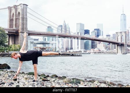 Mid adult man practicing yoga on riverside en avant du pont de Brooklyn, New York, USA Banque D'Images