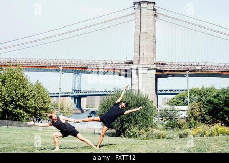 Deux hommes de vous pencher sur le côté en position de maintien de pied de yoga en face du pont de Brooklyn, New York, USA Banque D'Images