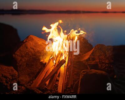 Feu et flammes de feu atmosphérique sur la plage le soir. Encore de l'eau du lac sur le fond avec des couleurs chaudes des soleils Banque D'Images