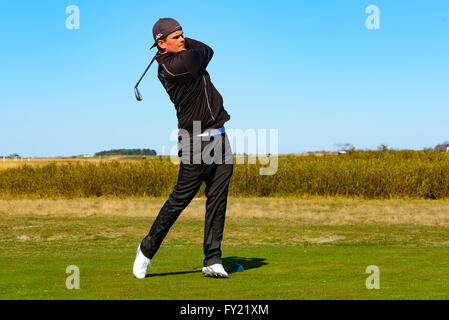 Skanor, Suède - 11 Avril 2016 : jeunes adultes mâles golfeur à la fin de son swing. Il est tous vêtus de noir et portent une casquette. Banque D'Images