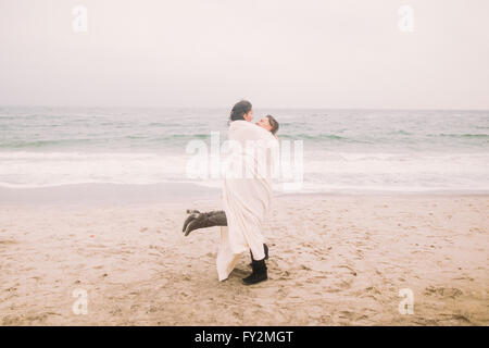 Heureux jeune homme détient sur les mains de sa charmante petite amie enveloppé dans une couverture blanche. Fond de mer d'hiver Banque D'Images