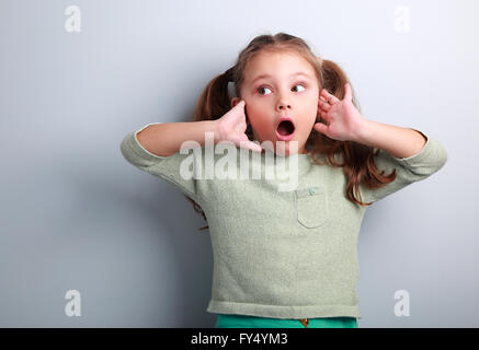 Surprenant kid girl with long hair avec bouche ouverte et la main près face à la recherche sur fond bleu copy space Banque D'Images