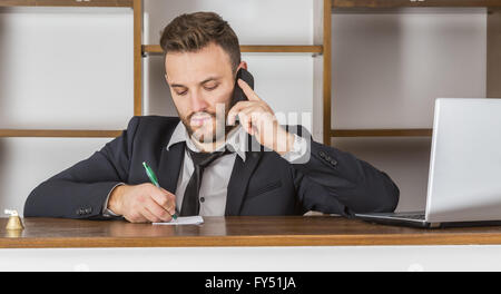 Portrait d'un réceptionniste prenant l'avis sur un morceau de papier lorsqu'il est au téléphone à son bureau dans une petite auberge. Banque D'Images