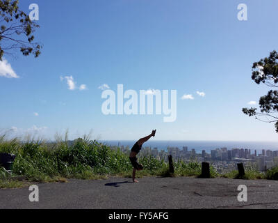 Homme portant un t-shirt, short et chaussons en caoutchouc Handstanding avec un dos courbé à Tantalus lookout point avec la ville de Honol Banque D'Images