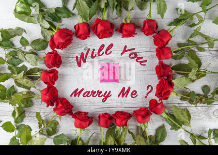 Allez-vous m'épouser ? Texte avec petit cadeau fort dans un cercle de roses rouges sur la table en bois blanc. Vue d'en haut. Banque D'Images