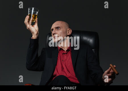 Handsome businessman avec verre de whisky et de cigare assis dans fauteuil de bureau sur fond noir Banque D'Images
