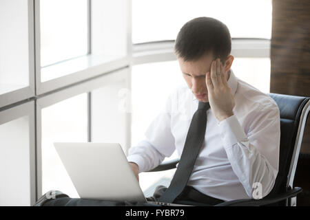 Beau jeune homme a souligné assis dans son bureau à l'aide d'ordinateur portable. Portrait d'affaires holding his head in hands Banque D'Images