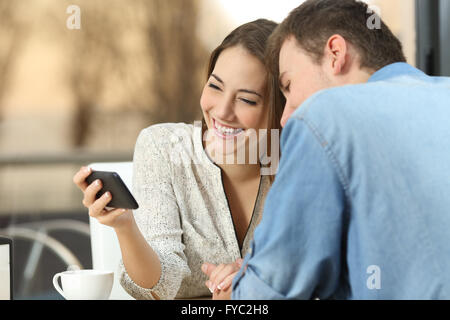 Happy casual couple rire et regarder des vidéos en streaming en ligne ensemble dans un smart phone générique dans un bar avec une ambiance chaleureuse Banque D'Images