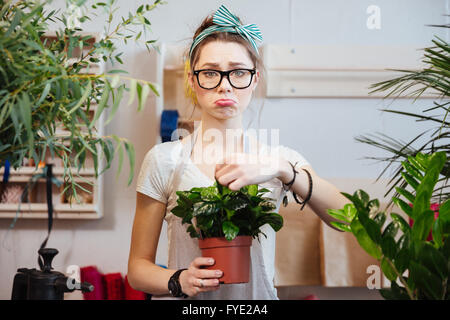 Triste belle jeune femme fleuriste à verres debout et tenant dans la plante verte en pot de fleurs Banque D'Images