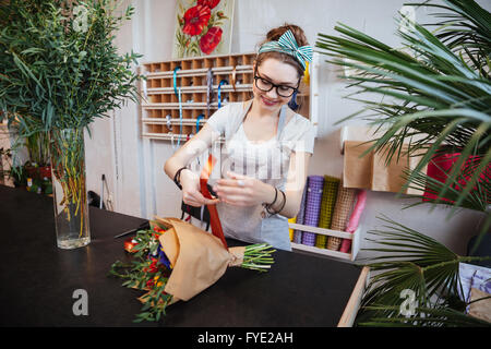 Smiling jolie jeune femme debout et faire fleuriste bouquet de fleurs avec ruban rouge Banque D'Images