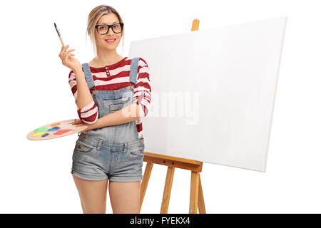 Young blonde woman holding a paintbrush devant une toile blanche isolé sur fond blanc Banque D'Images