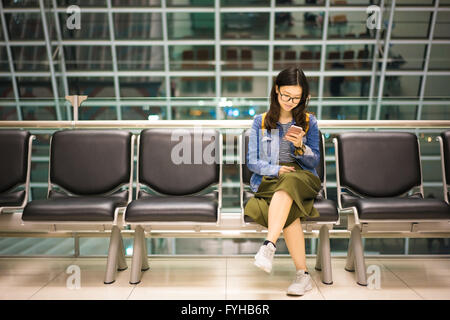 Belle fille asiatique siégeant seul, souriant à smartphone, en attendant l'embarquement, conceptuel de l'avion Banque D'Images