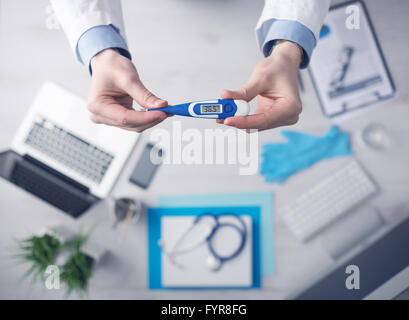 Médecin Mesure de la température et la tenue d'un thermomètre numérique, avec de l'équipement médical de bureau et l'ordinateur sur arrière-plan, top v Banque D'Images