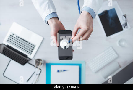 Médecin contrôle de pulsation sur téléphone mobile à l'aide d'un stéthoscope, desktop avec de l'équipement médical sur arrière-plan, medical app concept Banque D'Images