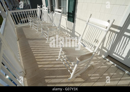 Une rangée de quatre utilisés et survécu à des chaises à bascule en bois peint blanc sur un porche sale au lever du soleil sur une journée ensoleillée. Banque D'Images