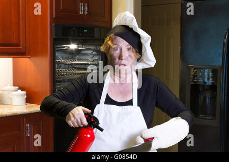 Mature Woman standing dans une cuisine avec visage roussis holding pan et extincteur avec chefs hat et un tablier. Banque D'Images