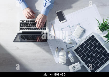 L'homme travaillant à son bureau sur un ordinateur portable avec l'économie d'énergie lampes CFL, un panneau solaire et un projet de maison, vue du dessus Banque D'Images