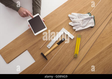 Les mains de charpentier à l'aide d'une tablette numérique et l'installation d'un parquet en bois avec outils de travail tout autour, vue du dessus Banque D'Images