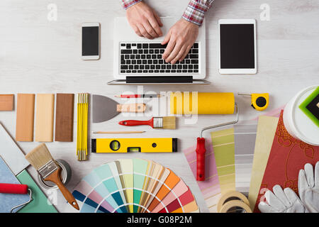 Les mains de professionnel de la décoration à son bureau de travail et de la saisie sur un ordinateur portable et des couleurs du nuancier, rouleaux de peinture et des outils sur la table de travail Banque D'Images