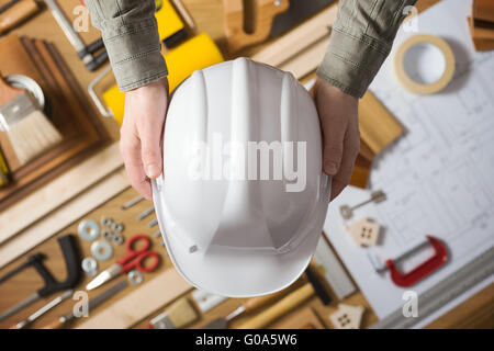 Mains tenant un casque de sécurité de protection contre une table de travail avec du matériel et des outils de construction, vue du dessus Banque D'Images