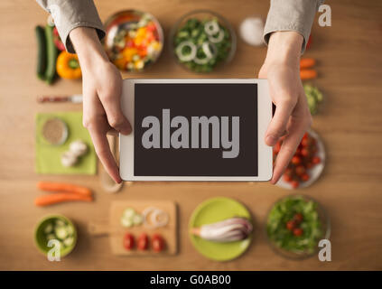 Mains tenant un écran tactile numérique avec des légumes frais et des ustensiles de cuisine sur l'arrière-plan, Vue de dessus Banque D'Images