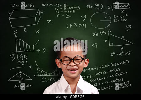 Petit Génie étudiant asiatique garçon avec des lunettes smiling over green chalkboard avec équivalents mathématiques écrit dessus Banque D'Images