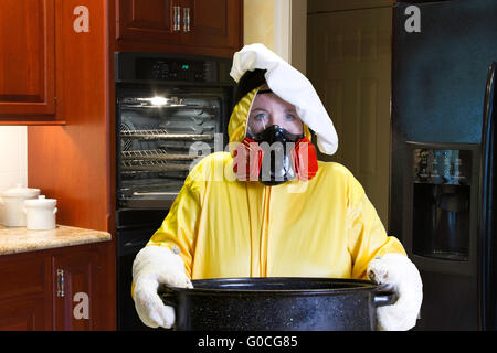 Mature femme portant une combinaison HazMat debout dans une cuisine avec visage roussis holding rôtissoire avec chefs hat. Banque D'Images