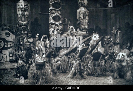 Les Kwakiutl ou Qagyuhi danseurs portant des masques et costumes lors d'un potlatch traditionnel 1914 en Colombie-Britannique, Canada. Le chef à l'extrême gauche est titulaire d'un personnel de l'Orateur. Trois mâts totémiques en arrière-plan. Banque D'Images