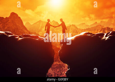 Deux hommes se serrent la main au-dessus du gouffre entre deux montagnes rocheuses au coucher du soleil. Business Banque D'Images