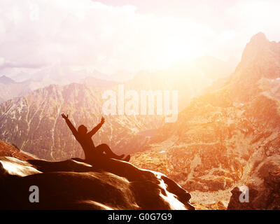 Un homme heureux assis sur la crête d'une montagne avec les mains levées en admirant une vue imprenable au coucher du soleil Banque D'Images