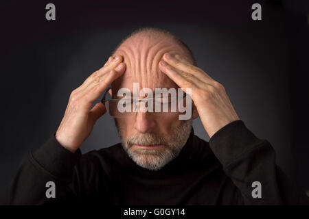 Fatigué - 60 ans homme avec une barbe et des lunettes à masser son front - un très gros plan sur un fond noir Banque D'Images