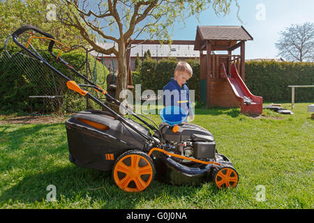 Mignon, petit garçon qui tond la pelouse. boy inspecte la tondeuse, avant de conduire ou de l'huile du moteur gauche. Banque D'Images